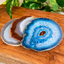 Load image into Gallery viewer, "Three Agate Slices stacked upon one another on a wooden plank with green foliage in the background," - Down to Earth.