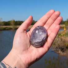 Load image into Gallery viewer, "Open hand holding a large Amethyst palm stone with a pond and green foliage in the background," - Down to Earth.