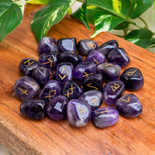 Load image into Gallery viewer, "Purple stones with runic symbols on a wooden surface with green leaves in the background," - Down to Earth.