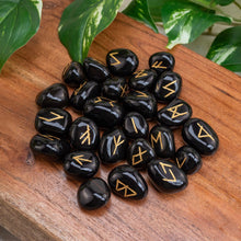 Load image into Gallery viewer, "Black Obsidian Rune Set on a wooden base with green foliage in the background," - Down to Earth.