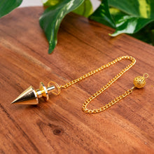 Load image into Gallery viewer, "Brass Pendulum that measures approximately 8.5 inches long on a wooden plank with green leaves in the background," - Down to Earth.