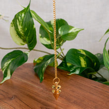 Load image into Gallery viewer, "Hanging Brass Pendulum hovering over a wooden base with green foliage in the background," - Down to Earth.