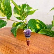 Load image into Gallery viewer, "Colorful crystal pendulum on a wooden surface with green leaves in the background," - Down to Earth.