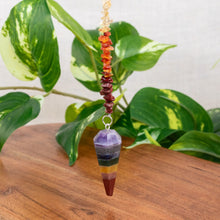 Load image into Gallery viewer, "Colorful crystal pendulum on a wooden surface with green leaves in the background," - Down to Earth.