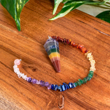 Load image into Gallery viewer, "Seven Chakra Pendulum with a crystal chip chain on a wooden plank with greenery in the background," - Down to Earth.
