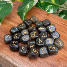 Load image into Gallery viewer, "Labradorite Rune Set with gold emblems engraved into the tumbled stones on a wooden plank with green foliage in the background," - Down to Earth.