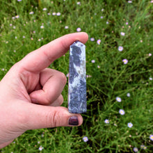 Load image into Gallery viewer, "Lepidolite tower pinched between a pointer finger and thumb with greenery in the background," - Down to Earth.