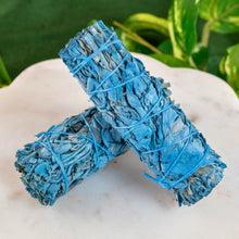 Load image into Gallery viewer, "Two blue Nag Champa White Sage smudge sticks on a white surface with greenery in the background," - Down to Earth.