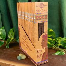 Load image into Gallery viewer, "Display case of Palo Santo Sacred Elements Incense Sticks with three green gemstones to the left on top of a wooden platform with foliage in the background," - Down to Earth.