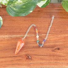 Load image into Gallery viewer, Premiere Carnelian Faceted Pendulum with 7 Chakra top down with plants on a style background - Down To Earth