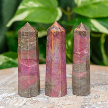 Load image into Gallery viewer, "Three Rhodonite Towers on a wooden base with green foliage in the background," - Down to Earth.