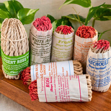 Load image into Gallery viewer, "Rope incense bundles in various colors and packaging on a wooden surface with plants in the background," - Down to Earth.