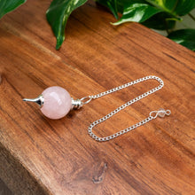 Load image into Gallery viewer, "Rose Quartz Sephoroton Pendulum on a wooden plank with green foliage in the background," - Down to Earth.