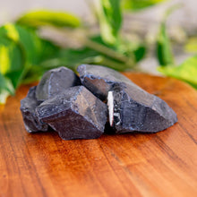 Load image into Gallery viewer, Raw blue goldstone crystals on a wooden surface with green leaves in the background