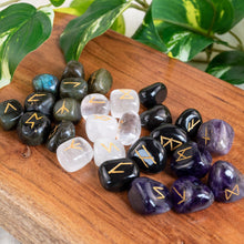 Load image into Gallery viewer, "Four sets of runes in Labradorite, Clear Quartz, Black Obsidian, and Amethyst, on a wooden plank with green foliage in the background," - Down to Earth.