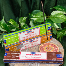 Load image into Gallery viewer, "Three packs of Satya Incense Sticks on a wooden plank next to a colorful vertical incense holder with green foliage in the background," - Down to Earth.