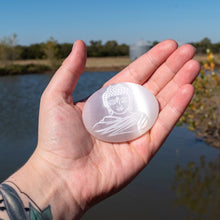 Load image into Gallery viewer, "Open hand holding a Selenite Buddha Palm Stone with a pond and green foliage in the background," - Down to Earth.