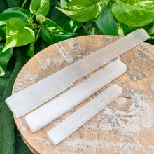 Load image into Gallery viewer, "Styled photograph of three sizes of Selenite wands on a wooden platform and green foliage in the background," - Down to Earth.