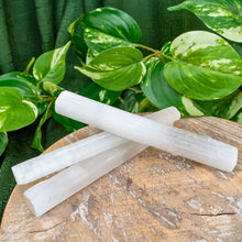 Load image into Gallery viewer, "Three medium Selenite wands on a wooden platform with green foliage in the background," - Down to Earth.