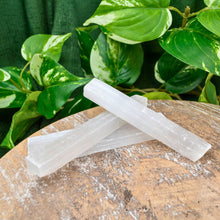 Load image into Gallery viewer, "Styled photo of three small Selenite wands on a wooden platform with green foliage in the background," - Down to Earth.