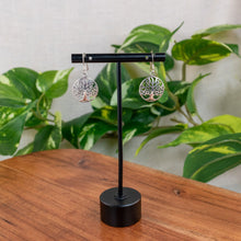 Load image into Gallery viewer, "Silver tree of life earrings on a black stand with green leaves in the background," - Down to Earth.