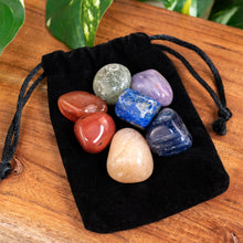 Load image into Gallery viewer, "Set of colorful tumbled stones on a black drawstring bag on a wooden surface with green leaves in the background," - Down to Earth.