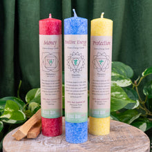 Load image into Gallery viewer, "Three Aloha Bay Chakra candles in the variations money, positive energy, and protection, on a wooden plank with green foliage in the background," - Down to Earth.