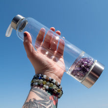 Load image into Gallery viewer, "Open palm holding an Amethyst crystal water bottle towards the sky with a clear blue sky in the background," - Down to Earth.