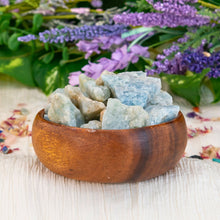 Load image into Gallery viewer, "A wooden bowl overflowing with raw aquamarine crystals with flowers in the background," - Down to Earth.