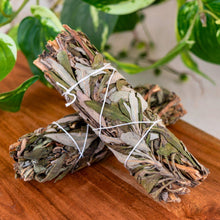 Load image into Gallery viewer, "Two bundles of black sage tied together on a wooden surface with green leaves in the background," - Down to Earth.