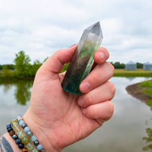 Load image into Gallery viewer, "Hand holding a Fluorite tower with a pond and greenery in the background," - Down to Earth.