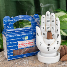 Load image into Gallery viewer, "Single package of Nag Champa Satya Backflow Dhoop Cones next to a Palmistry Incense Burner on a wooden plank with green foliage in the background," - Down to Earth.