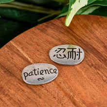 Load image into Gallery viewer, "Two silver oval pendants that say 'patience' in English on one side and in Kanji on the other on top of a wooden base with green foliage in the background," - Down to Earth.
