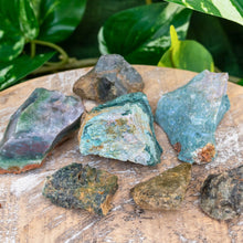 Load image into Gallery viewer, "Seven pieces of raw Sea Jasper on a wooden plank with green foliage in the background," - Down to Earth.