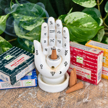 Load image into Gallery viewer, "Group of four packages of Satya Dhoop Cones with a Palmistry Incense Burner in the center with green foliage in the background," - Down to Earth.
