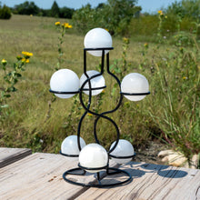 Load image into Gallery viewer, "Seven selenite spheres held by a metal stand with greenery in the background," - Down to Earth.