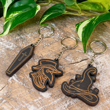 Load image into Gallery viewer, "Wooden Spooky Keychains on a wooden plank with green foliage in the background," - Down to Earth.