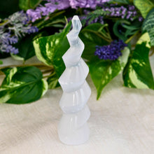 Load image into Gallery viewer, &quot;White selenite spiral crystal tower on a white cloth with green leaves and purple flowers in the background,&quot; - Down to Earth.
