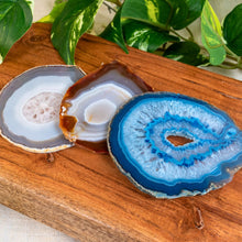 Load image into Gallery viewer, "Agate slices on a wooden surface with green leaves in the background," - Down to Earth.
