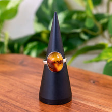 Load image into Gallery viewer, "Ring with amber stone on a black cone stand against a blurred green plant background," - Down to Earth.

