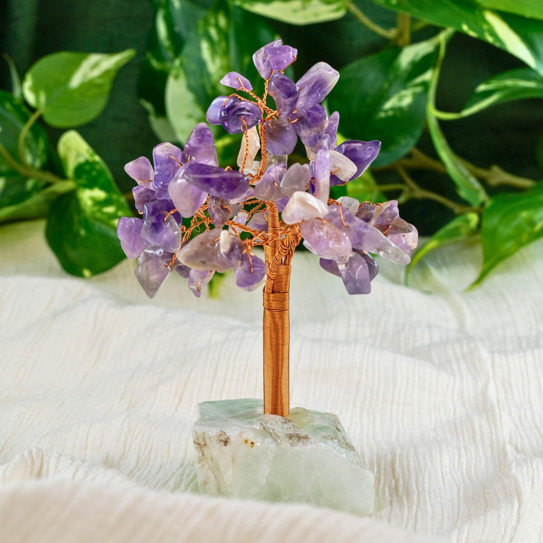 Decorative crystal tree with amethyst stones on a green base, surrounded by green leaves.