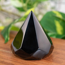 Load image into Gallery viewer, "Faceted Black Obsidian Chakra Extractor on a wooden plank with green foliage in the background," - Down to Earth.
