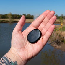 Load image into Gallery viewer, &quot;Open hand holding a Black Tourmaline Palm Stone with a pond and green foliage in the background,&quot; - Down to Earth.
