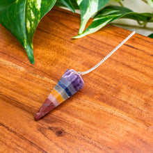 Load image into Gallery viewer, "Colorful Chakra Faceted Pendulum on a wooden surface with green leaves in the background," - Down to Earth.

