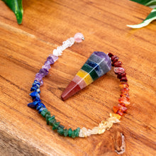 Load image into Gallery viewer, "Seven Chakra Pendulum with Crystal Chips on a wooden plank with green foliage in the background," - Down to Earth.
