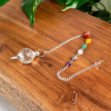 Load image into Gallery viewer, "Quartz Globe Pendulum with Chakra Chain on a wooden plank with green foliage in the background," - Down to Earth.
