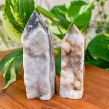 Load image into Gallery viewer, "Two crystal towers on a wooden surface with green leaves in the background," - Down to Earth.
