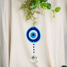 Load image into Gallery viewer, "Blue Evil Eye Suncatcher in front of a beige curtain with green foliage," - Down to Earth.
