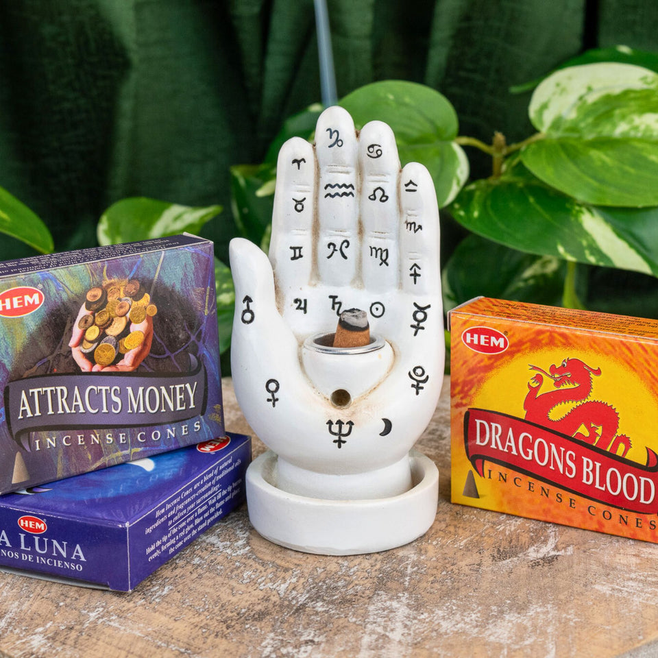 "Three packs of HEM Incense Cones surrounding a Palmistry Incense Burner with green foliage in the background," - Down to Earth.