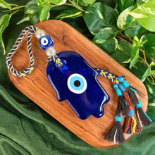 Load image into Gallery viewer, "Blue evil eye charm door hanger with tassels on a wooden board with green leaves in the background," - Down to Earth.
